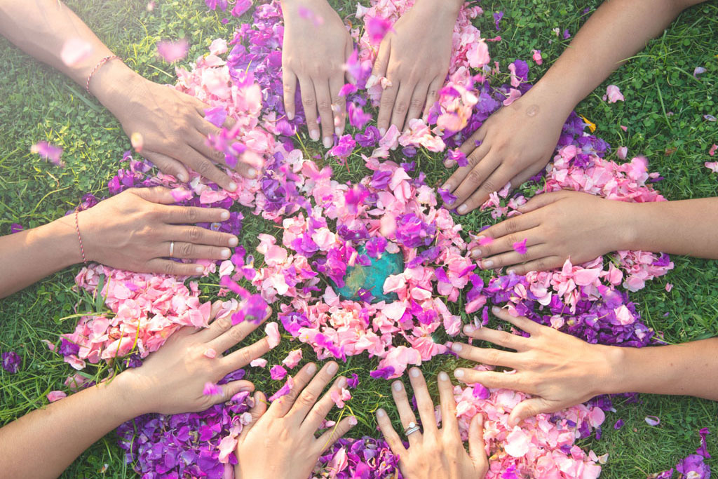 A circular arrangement of hands touching colorful flower petals in a mandala or danmala on green grass, with soft pink and purple hues blending together.