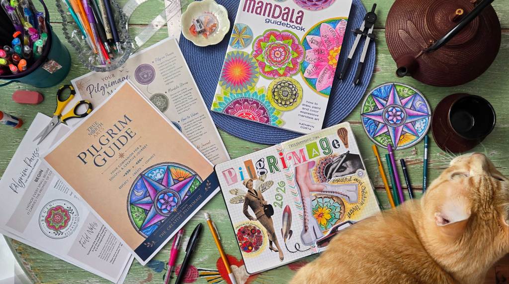 Table with an orange tabby, The Mandala Guidebook, journal, tea pot and cup, pages from the Pilgrim Guide, assorted drawing and coloring tools scattered and a mandala of a compass rose.